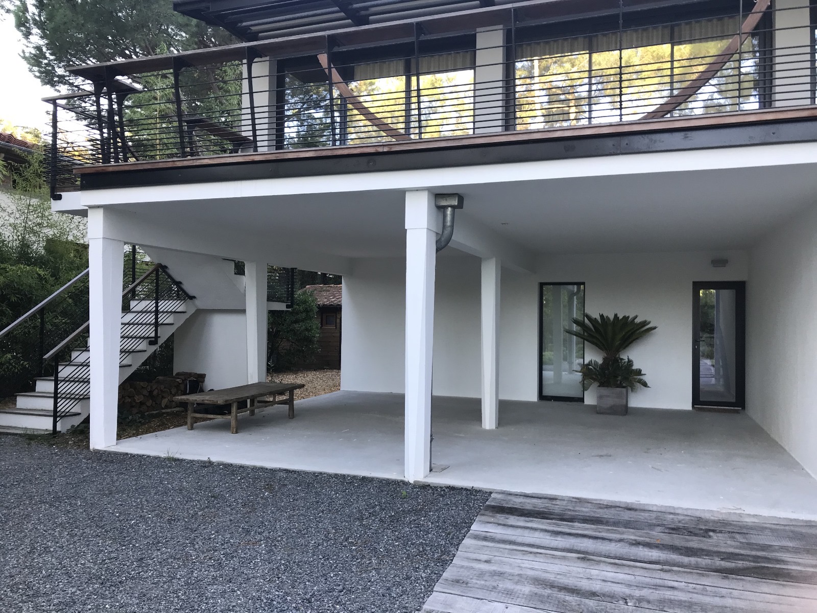 Architecture moderne sous un balcon, avec carport blanc, escalier extérieur noir, sol en gravier et une grande jardinière.