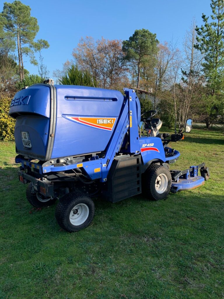Iseki SF450 : Tondeuse professionnelle avec bac de ramassage Tondeuse professionnelle Iseki SF450, de couleur bleue, avec bac de ramassage arrière, garée sur une pelouse verte sous un ciel bleu.