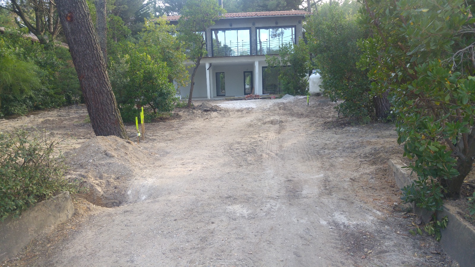 Chemin de terre montrant un chantier de construction moderne à deux étages avec grandes baies vitrées, entouré de végétation.