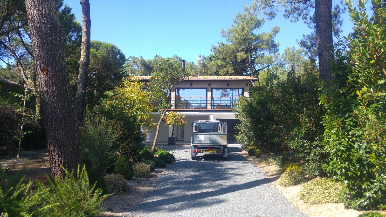 Maison bois moderne, pinède et allée gravier Moderne maison boisée avec balcon, accessible par une allée de gravier où une petite remorque immatriculée 3169-SR-33 est garée.